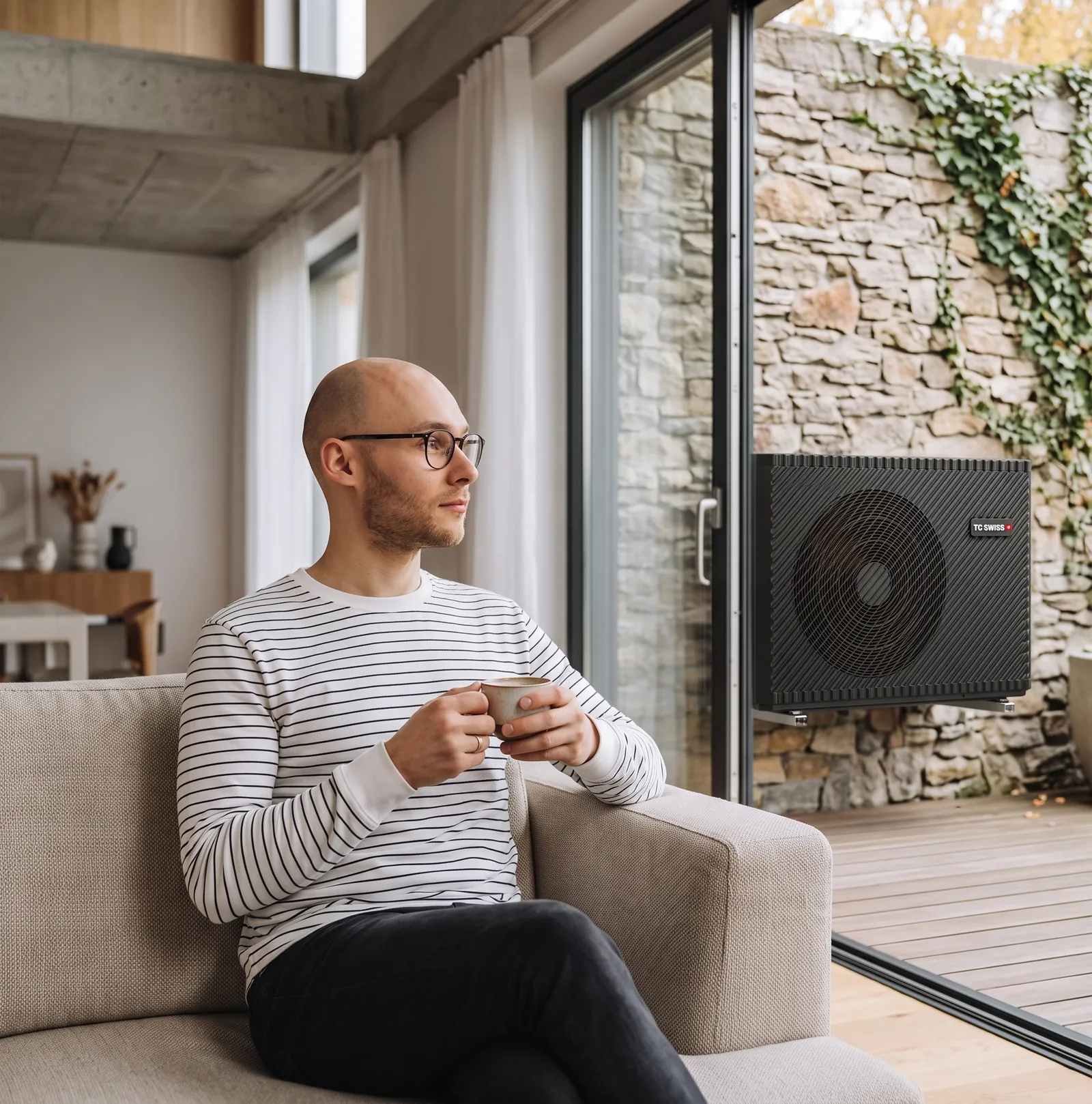 Bewoner met een kop koffie op de bank; buiten het raam hangt een TC Swiss warmtepomp